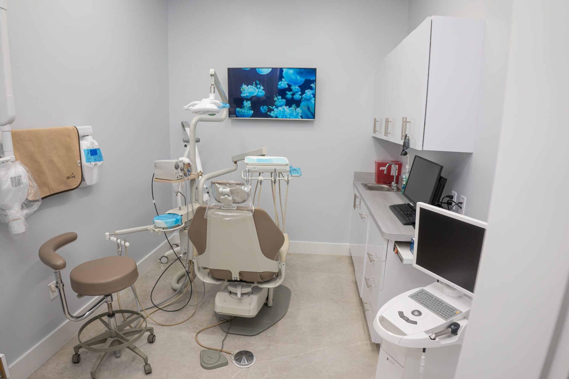 A well-equipped, modern dental operatory with a beige dental chair, digital monitors showing jellyfish, and professional cabinetry.