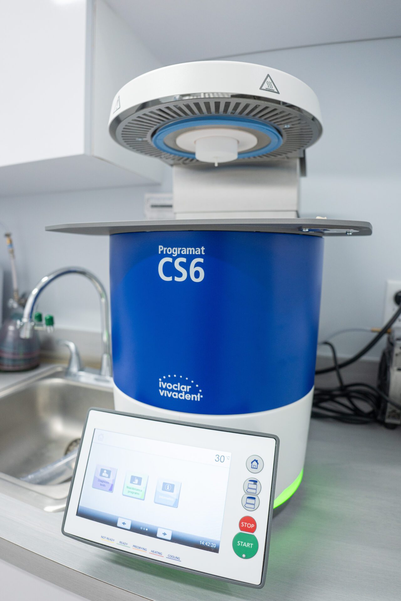 Close-up of a blue and white Ivoclar Vivadent Programat CS6 dental furnace with an open heating chamber and a digital touchscreen controller.