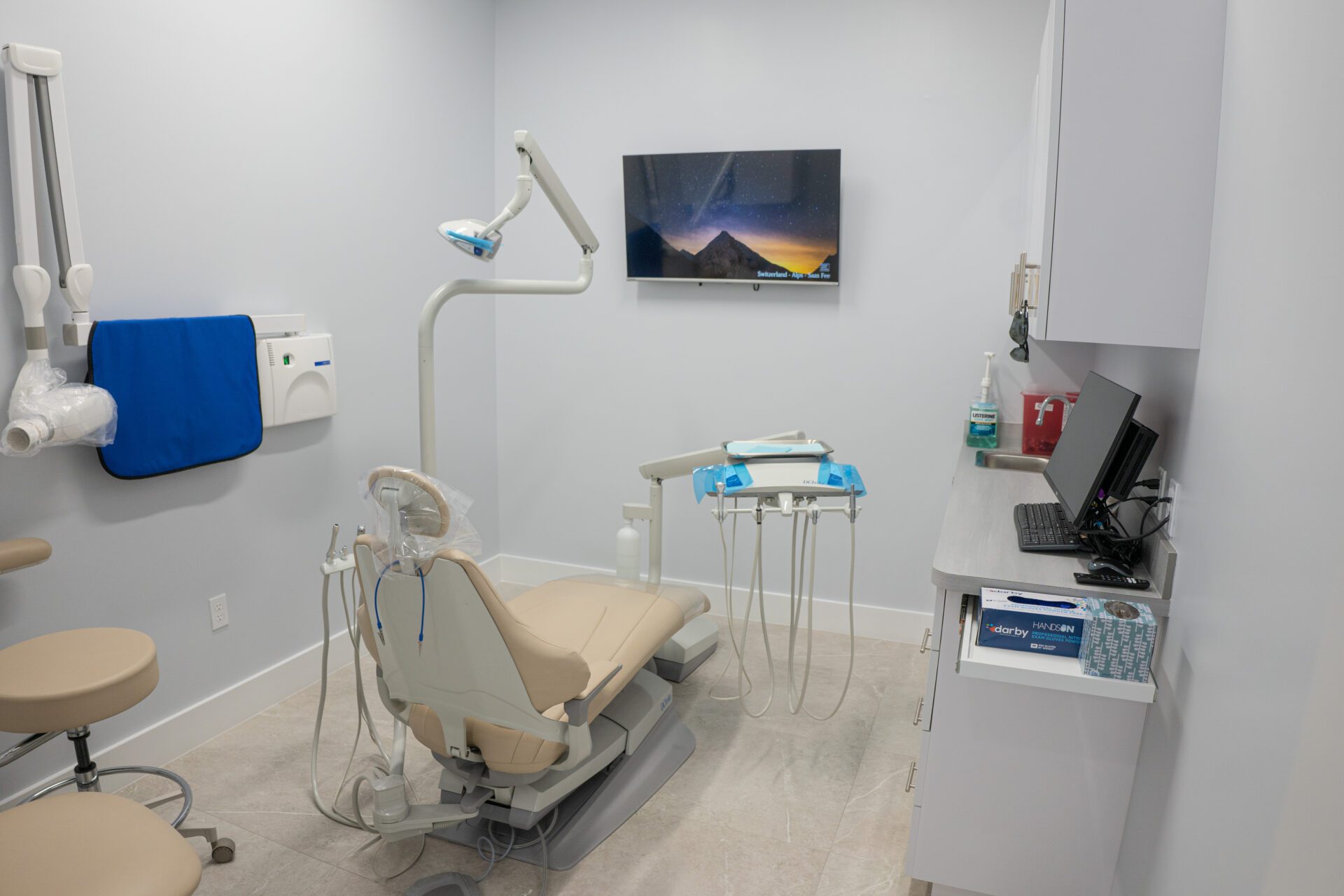 A clean, modern dental examination room featuring an adjustable beige chair, overhead exam light, wall-mounted X-ray machine, and a computer workstation.