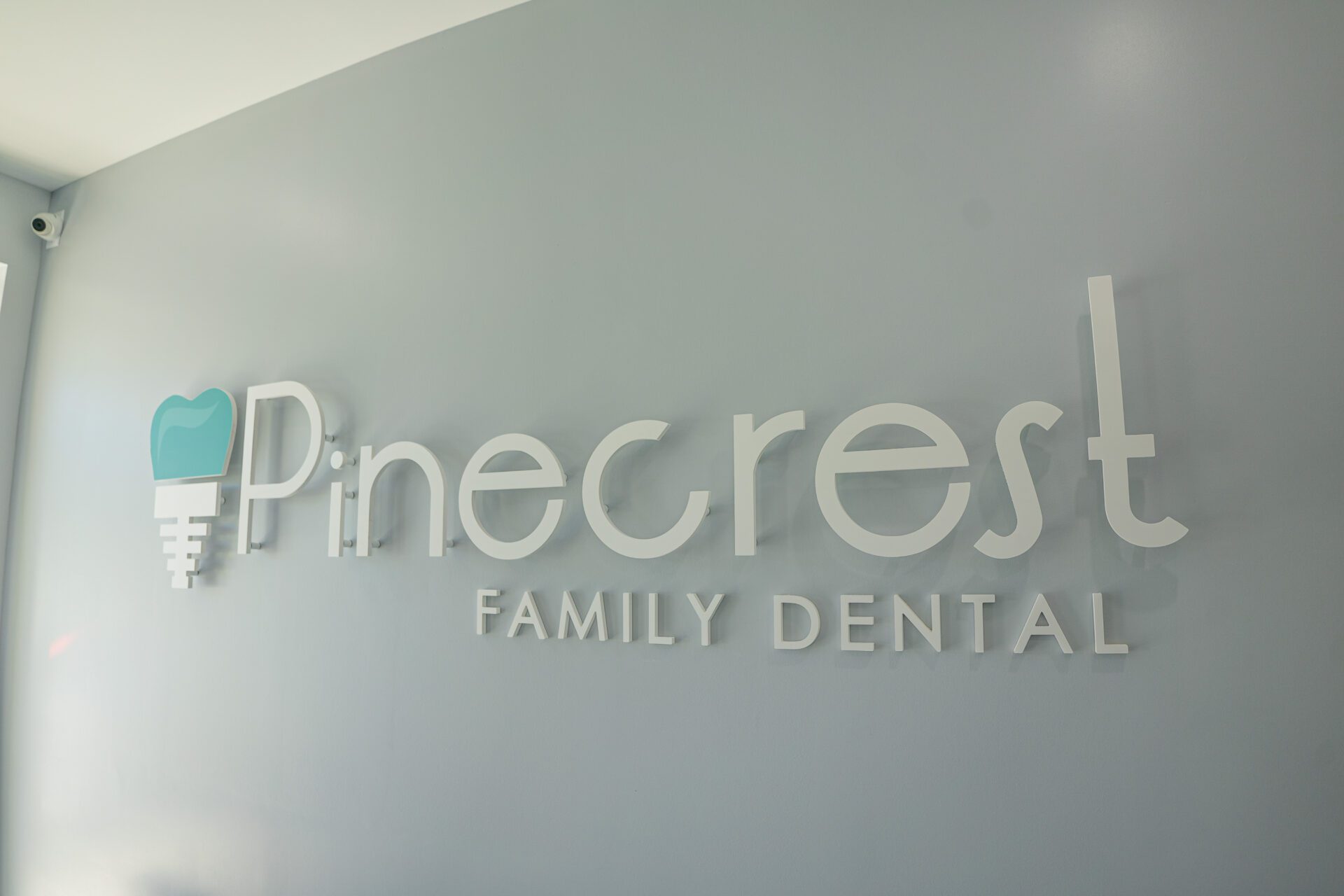 Modern wall-mounted 3D sign for Pinecrest Family Dental with a teal tooth logo on a grey wall.