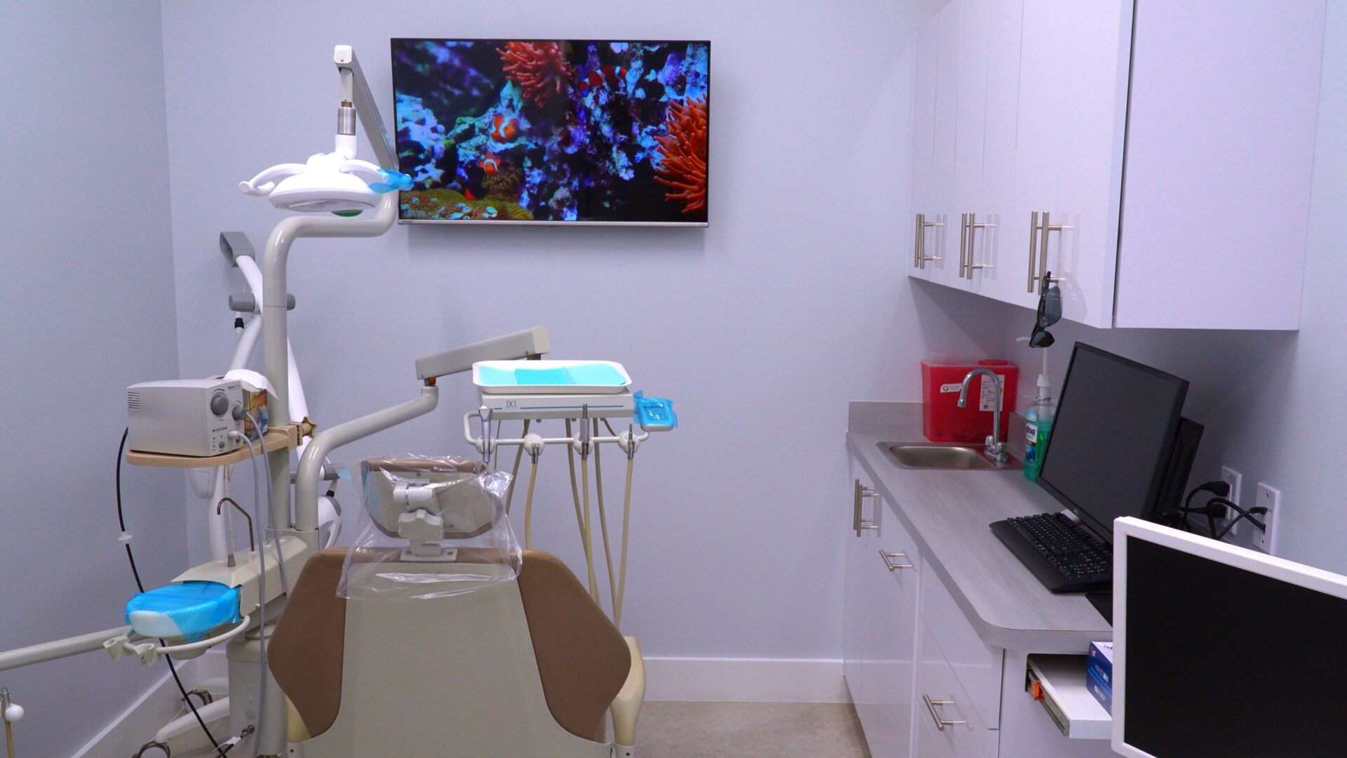 A high-tech dental operatory with a comfortable chair, specialized lighting, a delivery unit for dental tools, and a large wall-mounted monitor showing an underwater scene for patient relaxation.