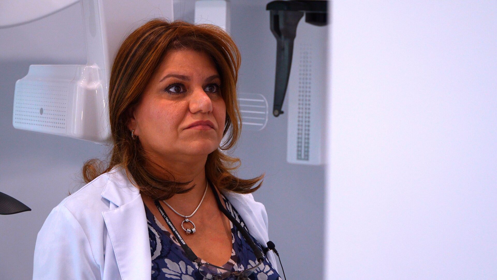 A professional female dentist wearing a white lab coat standing in front of high-tech dental imaging equipment in a clinic.