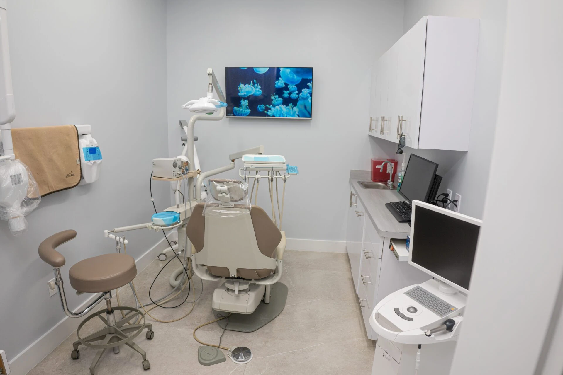 A well-equipped, modern dental operatory with a beige dental chair, digital monitors showing jellyfish, and professional cabinetry.