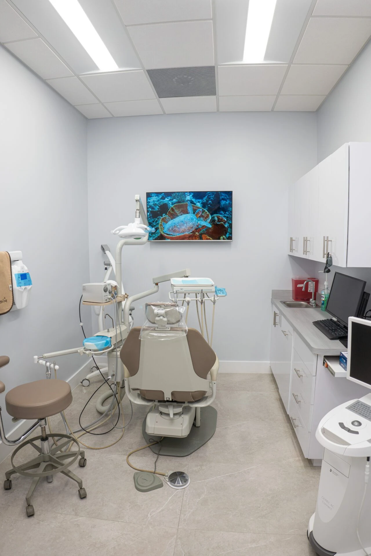 A clean, modern dental exam room featuring a tan patient chair, white cabinetry, high-tech dental equipment, and a large TV screen displaying a sea turtle.