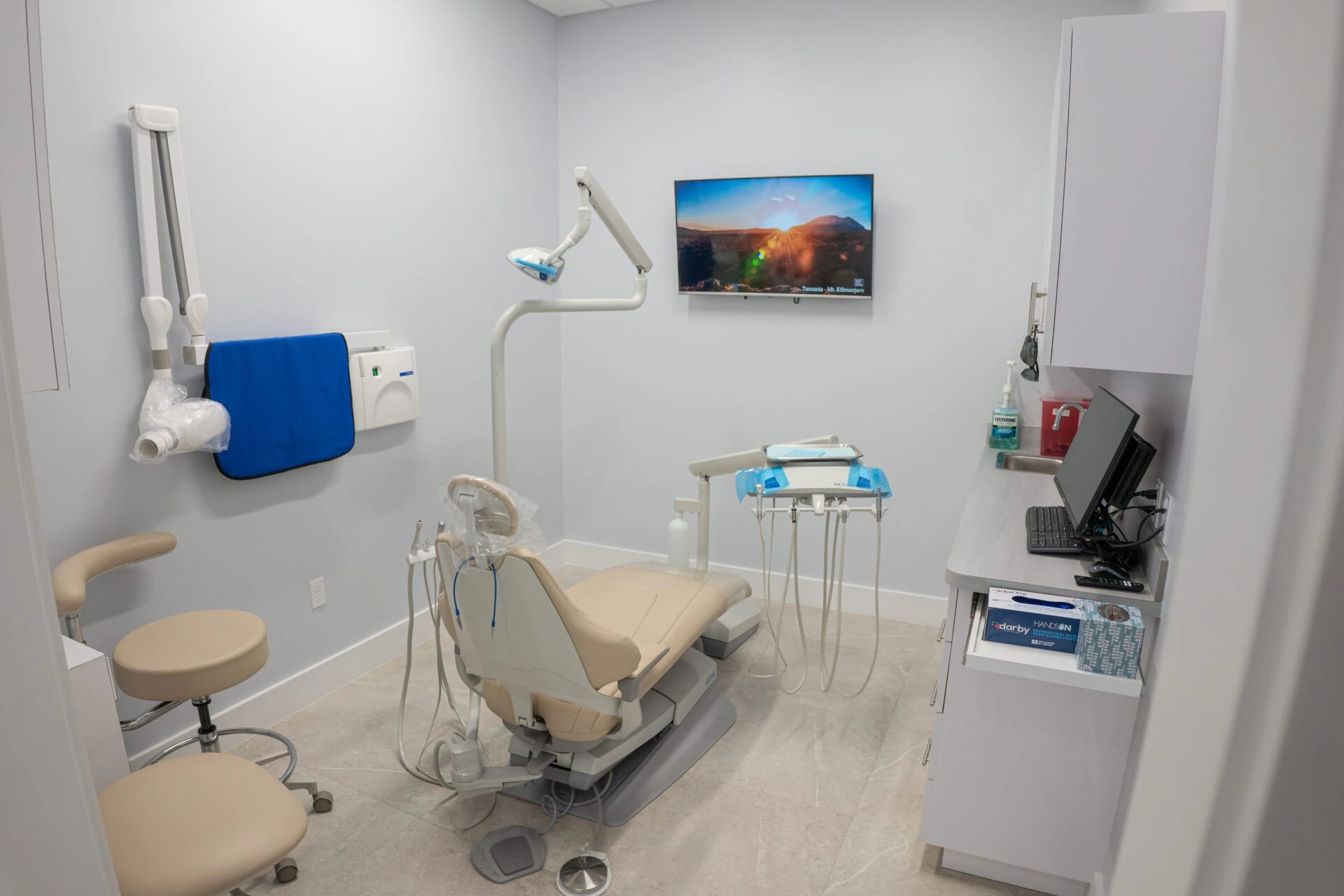Interior view of a modern dental treatment room featuring a tan leather dental chair, specialized medical equipment, an X-ray machine, and a wall-mounted TV for patient comfort.