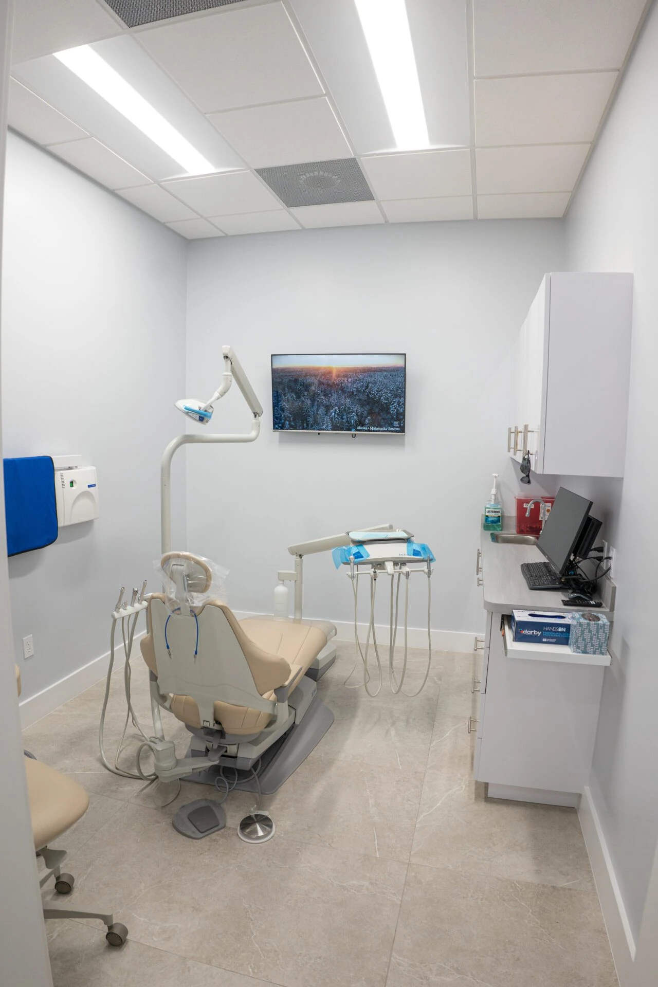 A modern dental examination room with a beige patient chair, advanced dental equipment, and a wall-mounted TV in a bright, clean office setting.