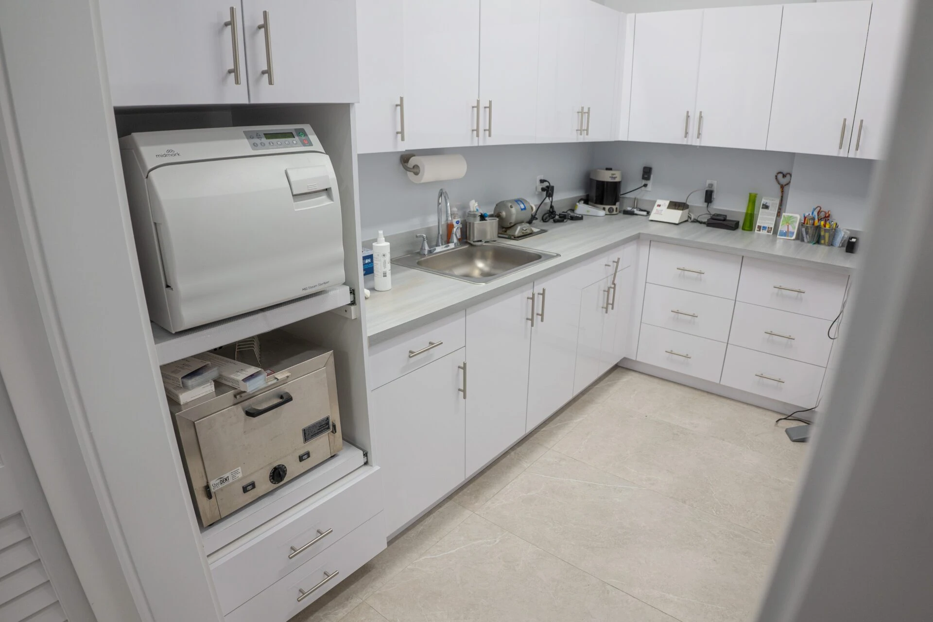 A clean, modern medical sterilization room featuring white cabinetry, grey countertops, a Midmark M11 autoclave, and a Steri-Dent dry heat sterilizer.