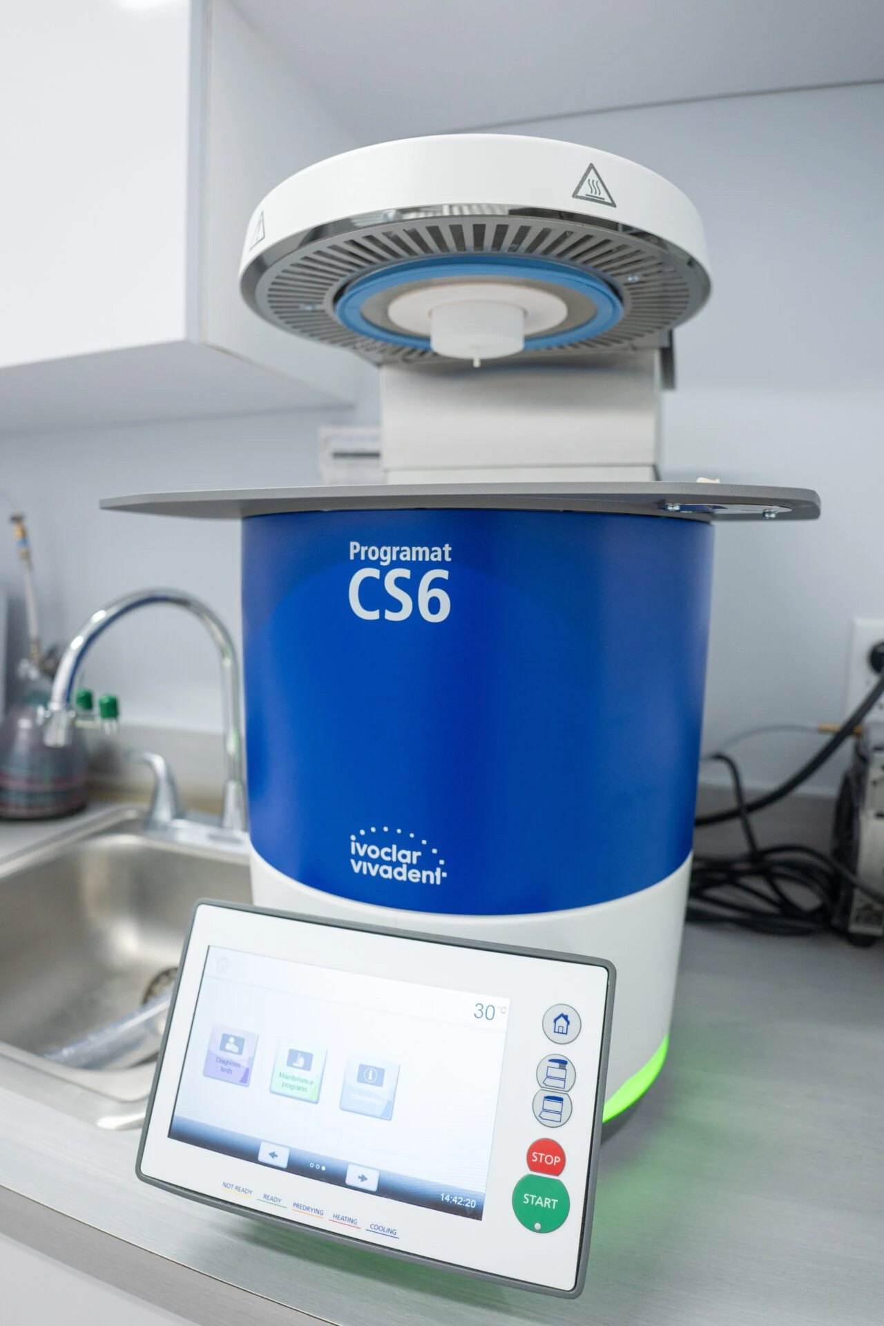 Close-up of a blue and white Ivoclar Vivadent Programat CS6 dental furnace with an open heating chamber and a digital touchscreen controller.