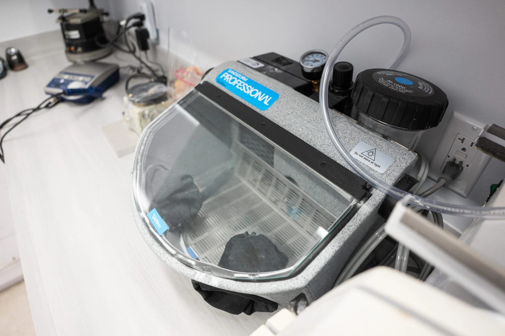 A Vaniman Sandstorm Professional abrasive sandblaster unit sitting on a white dental laboratory countertop with integrated gloves and a clear shield.