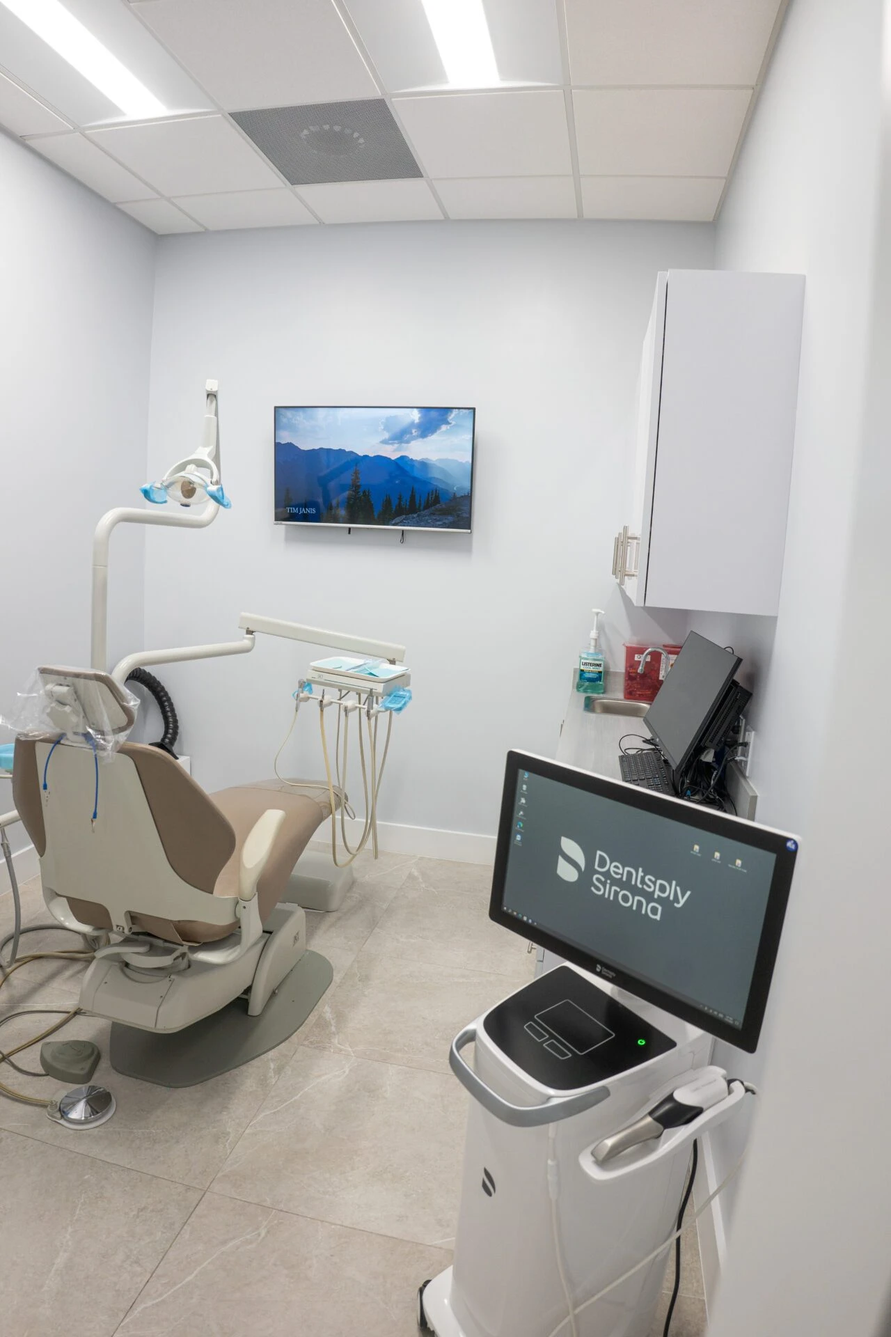 A clean, modern dental operatory featuring a tan dental chair, an overhead surgical light, and a Dentsply Sirona intraoral scanner on a mobile cart. A television on the wall displays a scenic mountain landscape.