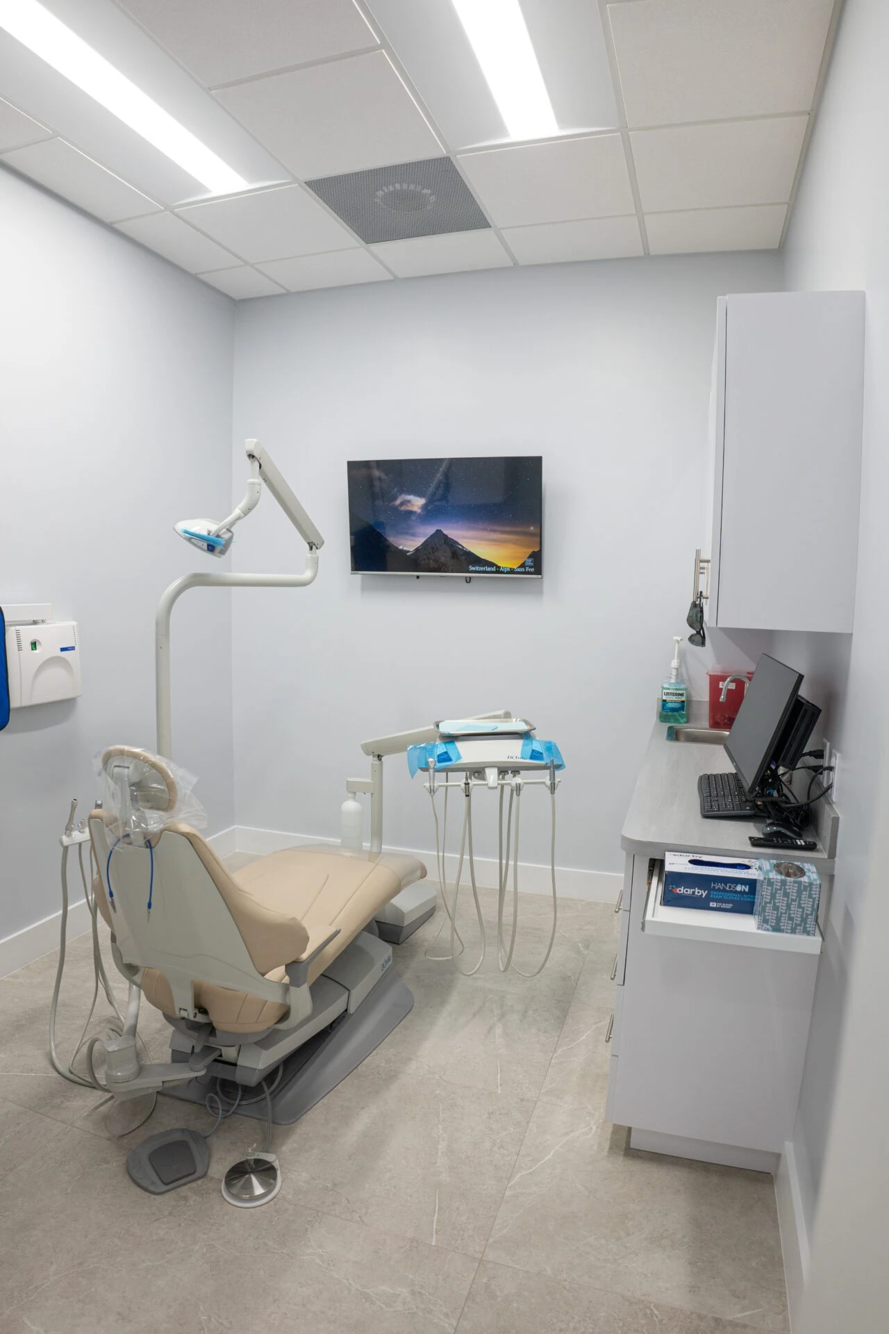 A clean and modern dental treatment room featuring a beige patient chair, advanced dental equipment, a wall-mounted TV, and a computer workstation.