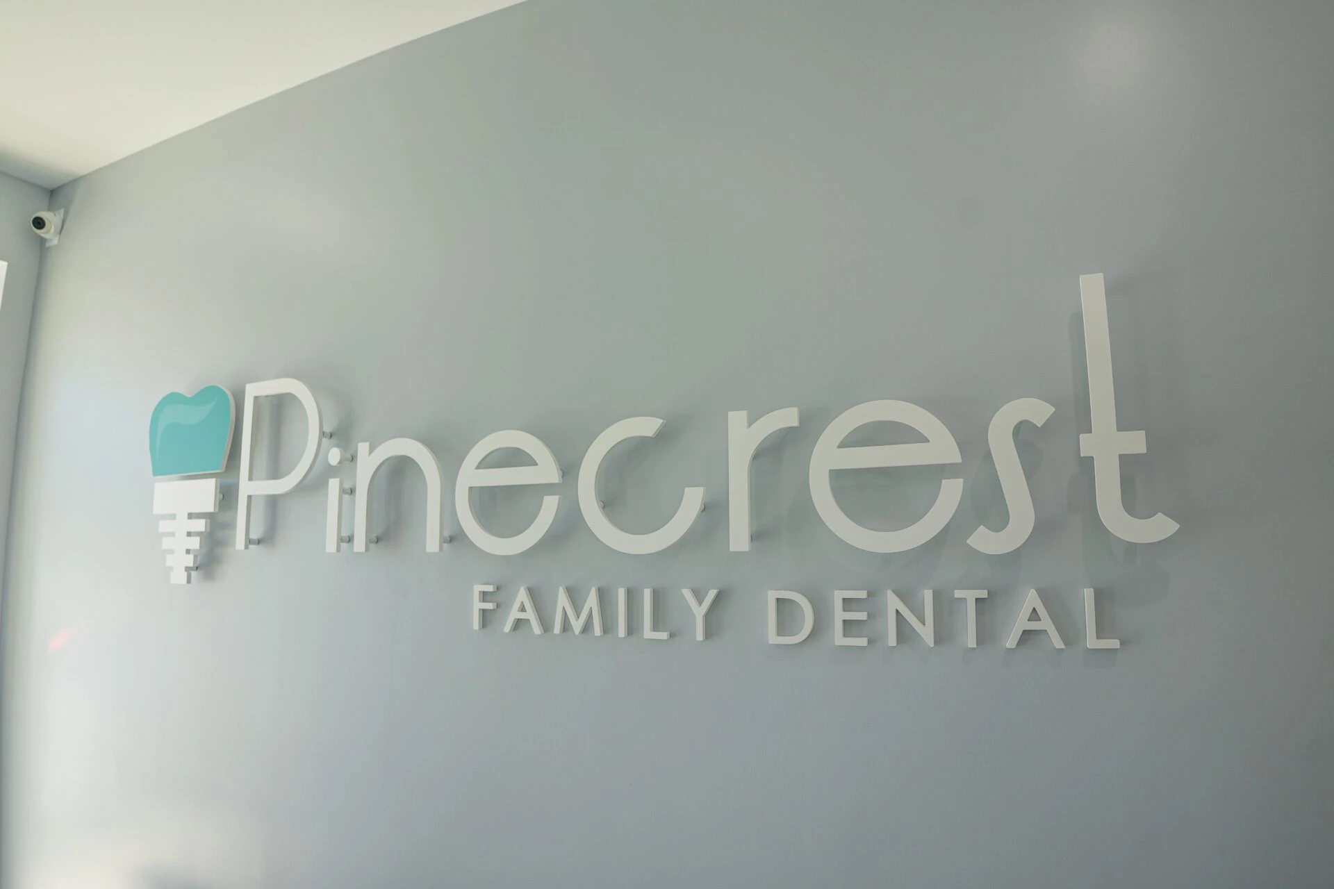 Modern wall-mounted 3D sign for Pinecrest Family Dental with a teal tooth logo on a grey wall.