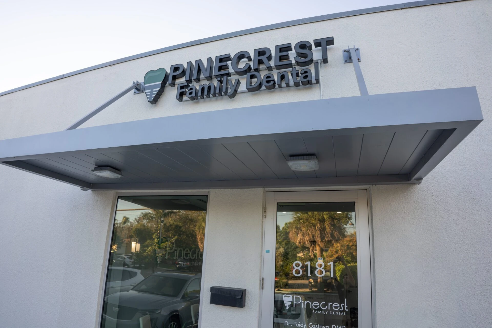 The modern exterior storefront of Pinecrest Family Dental, featuring a grey awning and large black signage above a glass entrance.