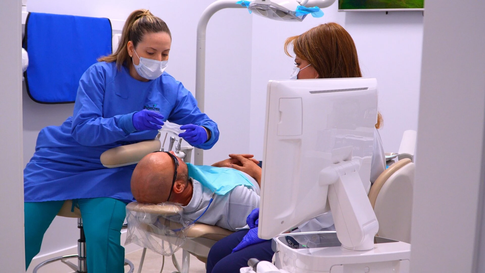 A dental assistant in blue scrubs prepares equipment for a patient lying in a dental chair while a dentist observes from a computer station.