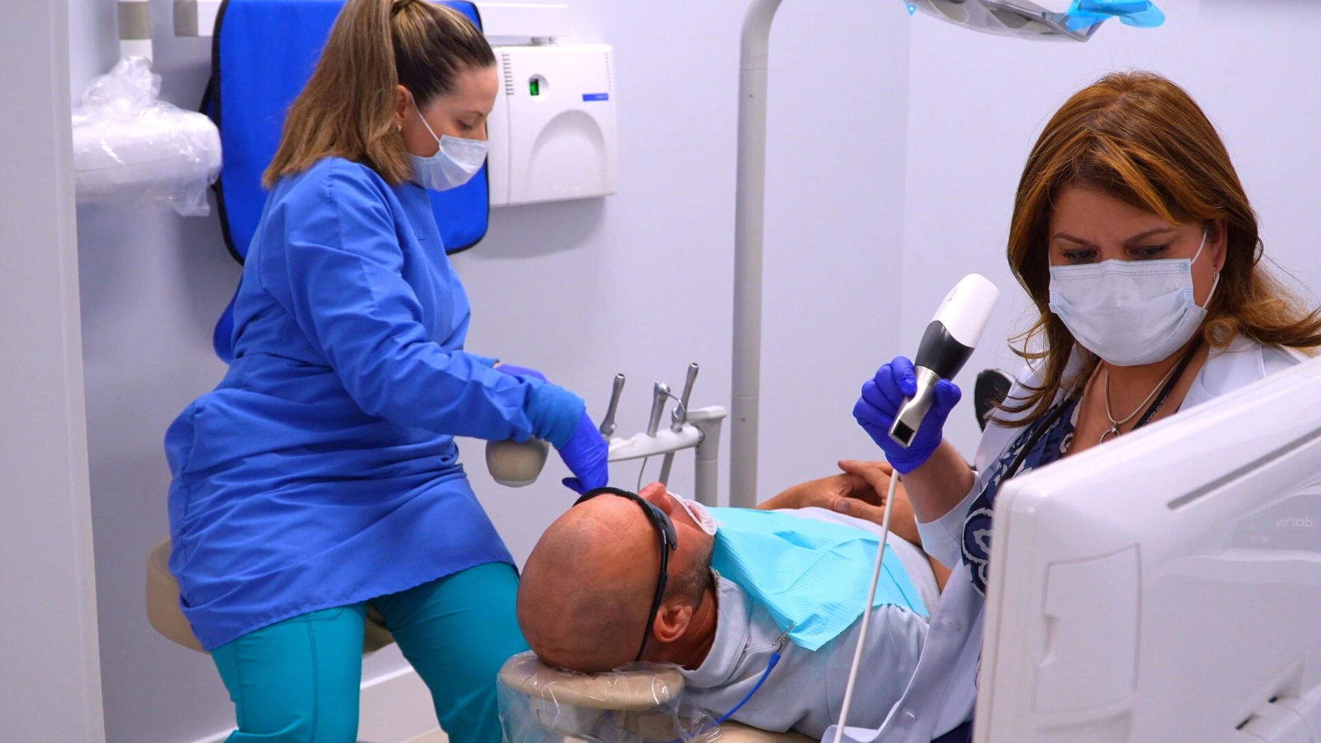 A female dentist wearing a mask and gloves uses a handheld intraoral scanner to create a digital impression of a male patient's teeth in a modern dental office.