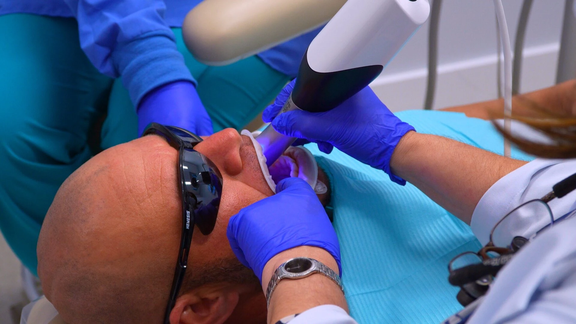 A dental professional wearing blue gloves uses a modern handheld intraoral scanner to capture digital impressions of a patient's teeth.