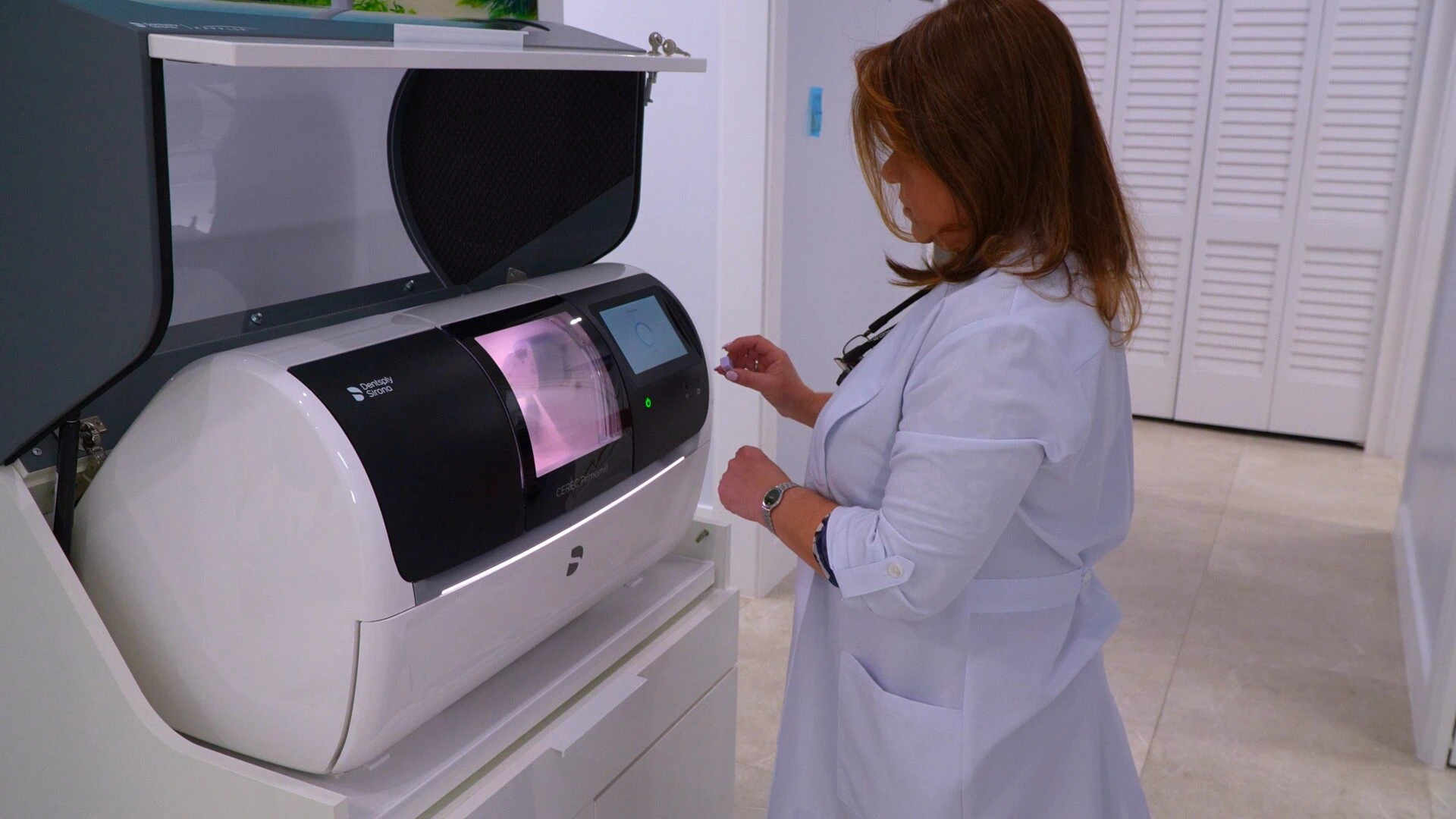 A dentist in a white lab coat using a CEREC Primemill machine to manufacture a dental crown in a clinic.