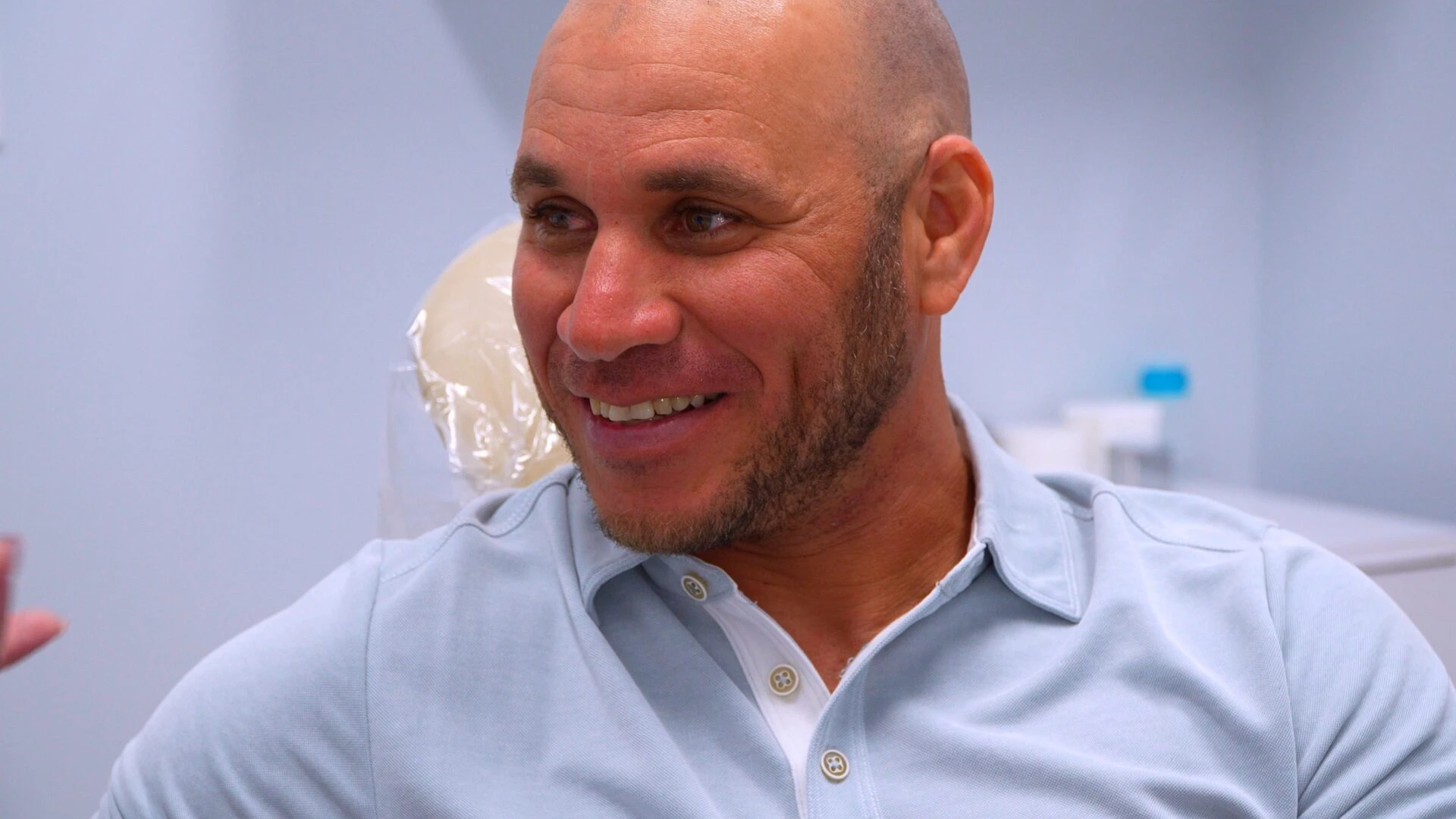 A smiling middle-aged man with a shaved head and beard sitting in a dental clinic setting.