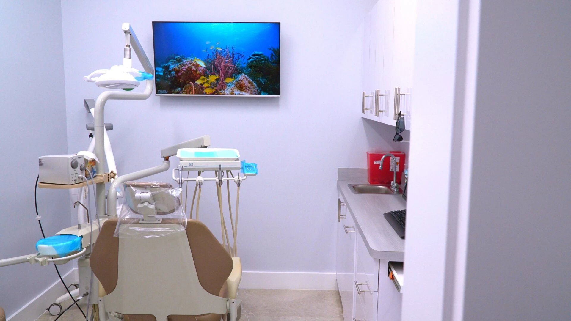 Interior view of a modern dental operatory featuring a tan patient chair, clinical workstation with a sink, and a large monitor displaying an underwater reef scene.