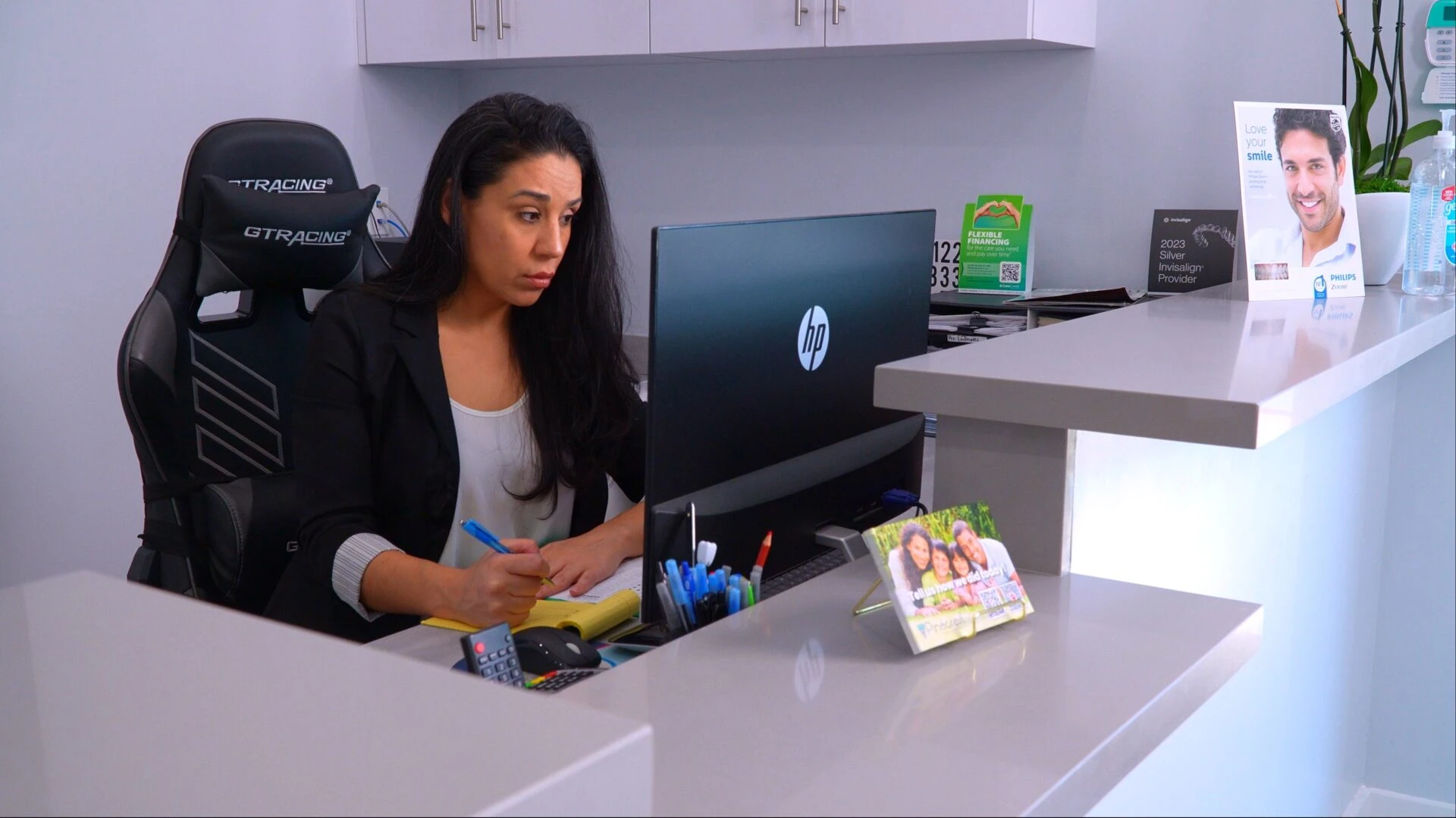 A professional woman in a black blazer working at a dental office reception desk, writing on a notepad while looking at a computer monitor.