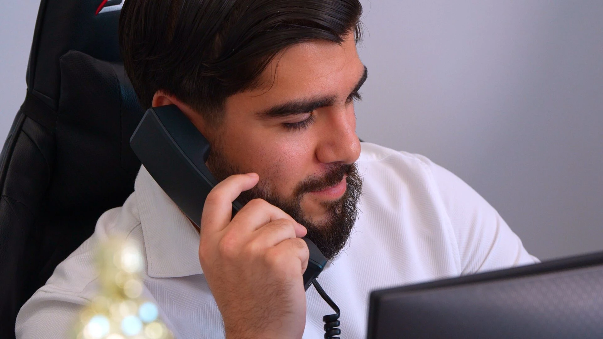 A young bearded businessman in a white shirt speaking into a black landline telephone receiver in an office setting.