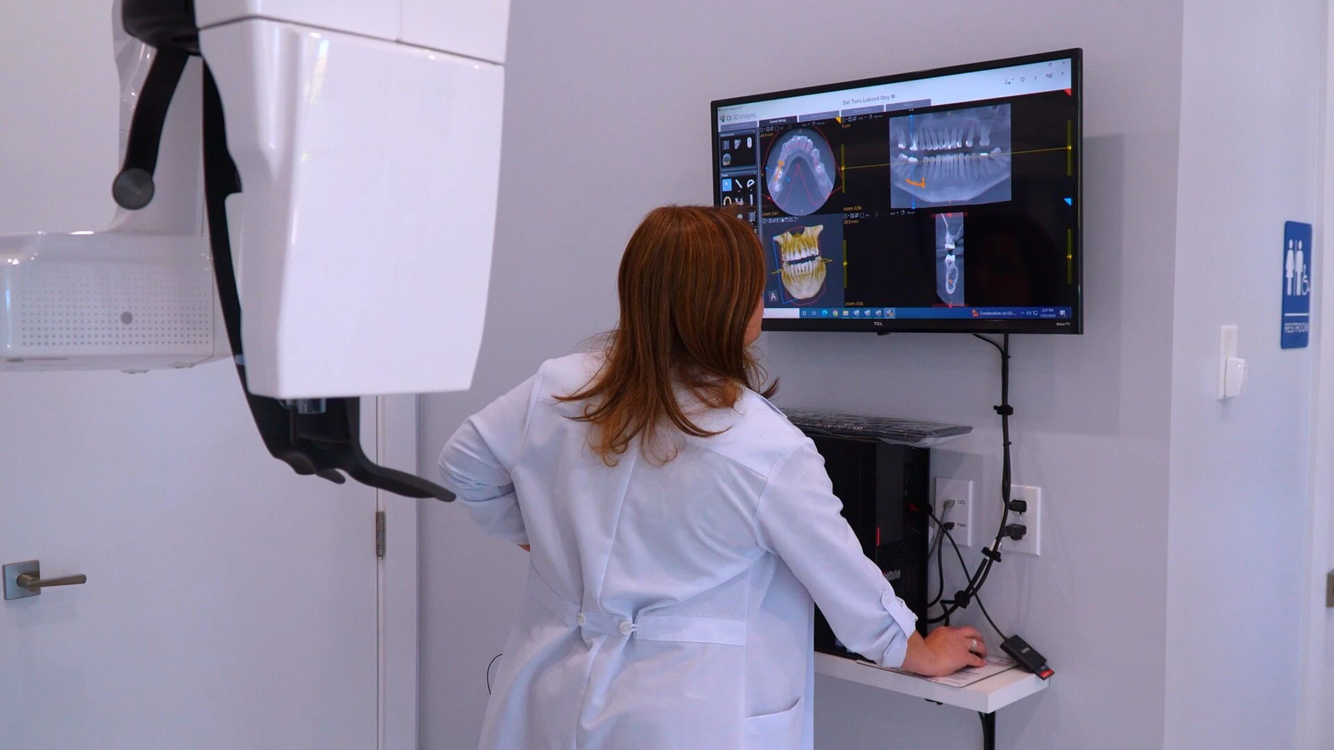 A dental professional in a white lab coat examines detailed 3D digital dental X-rays on a large wall-mounted monitor in a modern clinic.